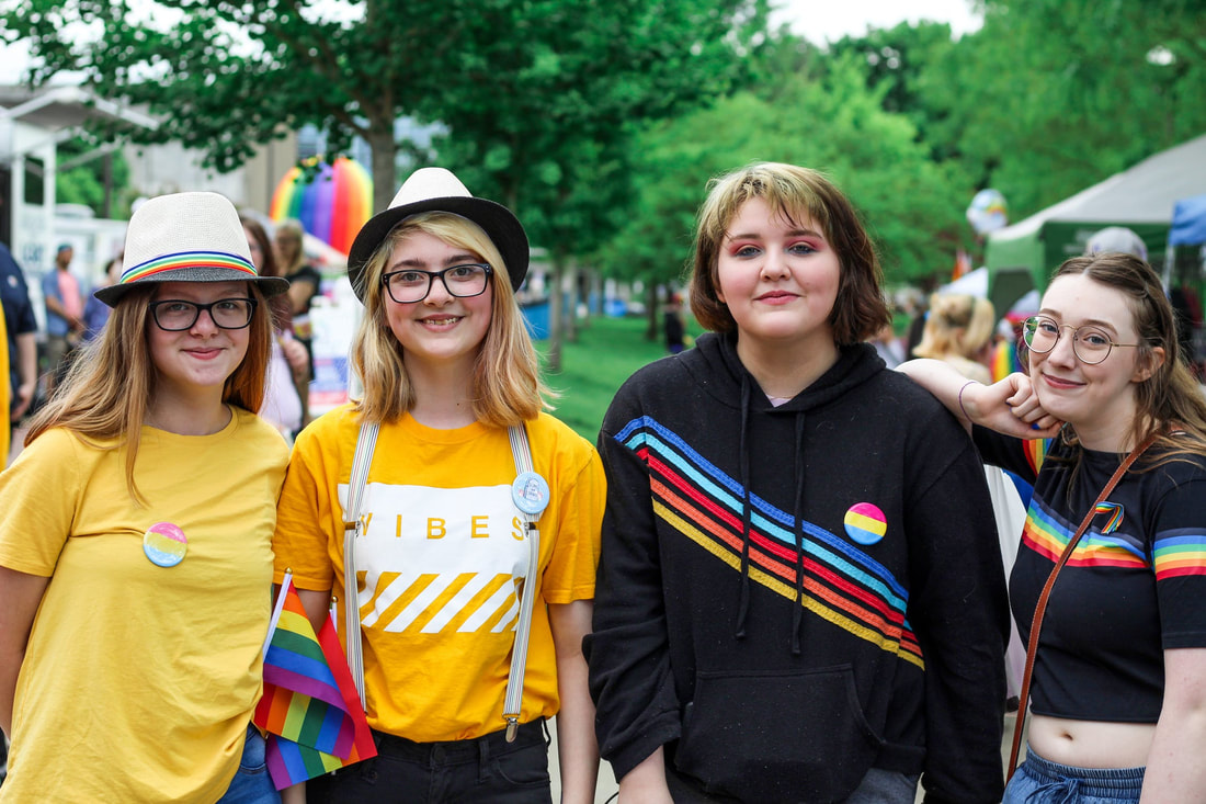 A group of four youths, smiling at the camera, decked out in various pride-related pins, hats, and shirts.